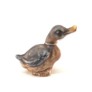 Puede incluir: Pequeña figura de pato de cerámica. El pato tiene un cuerpo marrón, alas grises y una cabeza oscura con pico amarillo. La figura es un objeto de colección, probablemente una pieza Wade, como indica el texto "WADE" en la base.