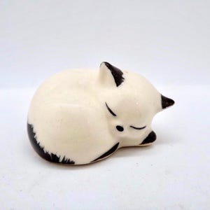 May include: A ceramic figurine of a sleeping fox. The fox is curled up, with its eyes closed. The figurine is primarily cream-colored with black accents on the ears, tail, and paws. The fox is resting on a white surface.