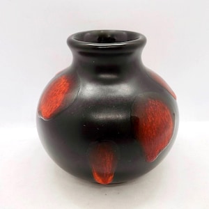 Könnte beinhalten: Kleine, runde schwarze Vase mit breiter Öffnung. Die Vase hat große, unregelmäßige rote Flecken auf ihrer Oberfläche. Die Vase hat eine glänzende Oberfläche und steht auf weißem Hintergrund. Die Keramikvase ist ein dekorativer Artikel.