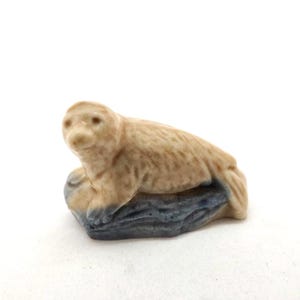 Op de afbeelding: Een kleine keramische figuur van een zeehond die op een rots rust. De zeehond is lichtbruin met een gladde textuur en de rots is een getextureerde blauwgrijs. De figuur staat tegen een witte achtergrond.