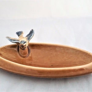 Puede incluir: Un plato de cerámica marrón con una figurita de pájaro en la parte superior. El pájaro está mirando hacia adelante con las alas extendidas. El plato tiene forma ovalada y un acabado liso y brillante.