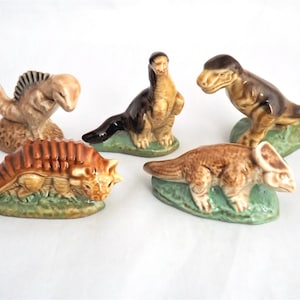 Puede incluir: Colección de cinco figuras de dinosaurios de cerámica en varias poses. Los dinosaurios están pintados en tonos marrones, beige y negro, con bases verdes. Las figuras incluyen un Spinosaurus, un Braquiosaurio, un Tyrannosaurus Rex, un Anquilosaurio y un Triceratops.