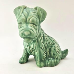 Puede incluir: Figura de perro de cerámica verde en posición sentada. La figura tiene una superficie texturizada y detalles, incluyendo orejas caídas y una cola corta. El perro es de color verde apagado.
