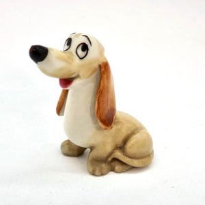 May include: A ceramic figurine of a cartoon dog in a seated position. The dog has a tan body, brown ears, and a black nose. Its eyes are wide and expressive, and its mouth is open, revealing a red tongue. The figurine is on a white background.