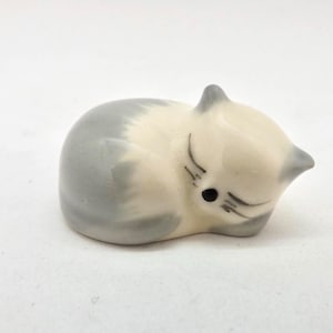 May include: A small, ceramic figurine of a sleeping cat. The cat is curled up, with its head resting on its paws. The cat is primarily cream-colored with gray markings on its head and back. Black lines depict the cat's closed eyes and whiskers.