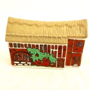 Può includere: Miniatura in ceramica di un mulino ad acqua con tetto di paglia. L'edificio è dipinto con pareti in mattoni rossi, una ruota idraulica e fogliame verde. Il tetto è beige e la base è bianca. Oggetto decorativo.
