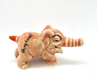 Könnte beinhalten: Eine rosa-orangefarbene Elefantenfigur aus Keramik. Der Elefant hat einen gestreiften Rüssel, ein lächelndes Gesicht und den Text "Oh, my head!" auf der Seite. Die Figur steht vor einem weißen Hintergrund.