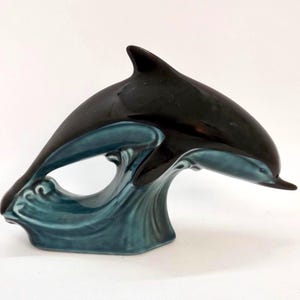 May include: A ceramic dolphin figurine in glossy black and teal blue. The dolphin is leaping over a wave, creating a dynamic and decorative piece. The figurine is a nautical-themed home decor item.