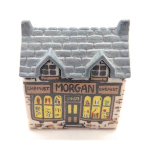 May include: A small ceramic chemist shop model with a grey tiled roof and two dormer windows. The shop front features the name "MORGAN" and "CHEMIST". The windows display various items, suggesting a pharmacy or drugstore.