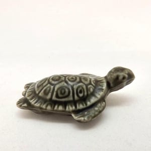 May include: A small ceramic turtle figurine in grey and green tones. The turtle features a detailed shell pattern, small head and legs. The figurine is set against a white backdrop.