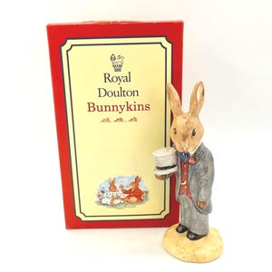 Puede incluir: Figura de Royal Doulton "Bunnykins" de un conejo con traje gris, sosteniendo un sombrero de copa. La figura está sobre una base beige, junto a una caja roja con el logotipo y texto de Bunnykins.