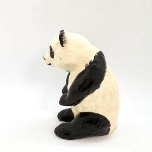 Könnte beinhalten: Eine Keramik-Panda-Figur in sitzender Position. Der Panda hat einen cremefarbenen Körper mit schwarzen Markierungen an Beinen, Armen sowie um Augen und Ohren. Die Figur ist ein dekorativer Artikel.