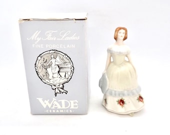Może przedstawiać: Porcelanowa figurka kobiety w długiej sukni, trzymającej kapelusz, obok pudełka. Na pudełku widnieje napis "My Fair Ladies FINE PORCELAIN" i "WADE - CERAMICS". Suknia figurki ma czerwone akcenty róż.