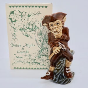 Puede incluir: Una figurita de cerámica marrón y verde de un duende sentado en una roca, sosteniendo un pico. La figurita es parte de una colección llamada "British Myths & Legends" de Wade.