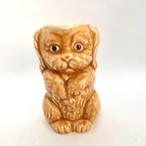 以下が含まれることがあります： 座っている姿勢の、薄茶色のセラミック子犬の置物。置物は濃い茶色の目を持ち、表面はざらざらしています。上部に小さな開口部があり、花瓶や容器として使用できる可能性があります。魅力的な装飾品です。
