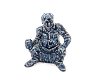 Könnte beinhalten: Blaue Keramikfigur einer sitzenden Person. Die Figur trägt ein Hemd mit Knöpfen und eine Hose. Die Figur steht vor einem weißen Hintergrund.