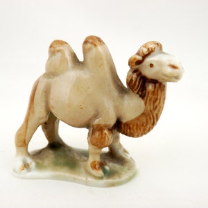 May include: A small ceramic figurine of a camel with two humps. The camel is light brown with a darker brown mane and tail. The figurine is standing on a green base.