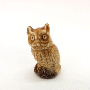 May include: A small, ceramic owl figurine in shades of brown and tan. The owl has detailed feather markings and large, round eyes. The figurine is perched on a small, textured base, and is a decorative item.