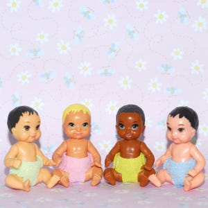 May include: Four baby dolls with different skin tones and hair colors. Each doll is wearing a diaper in a different pastel color: green, pink, yellow, and blue. The dolls are sitting on a pink floral background.
