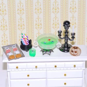 May include: Miniature Halloween-themed display featuring a cauldron with green liquid and eyeballs, a skull candelabra, a jack-o'-lantern, and a tray of cookies. A small green cup and a black cauldron with straws are also present. The display is set on a white dresser.