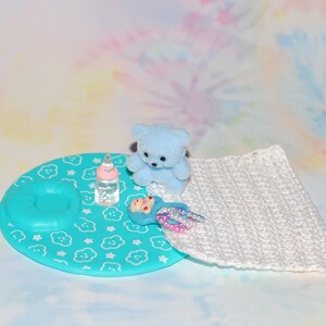 May include: A miniature baby-themed scene featuring a light blue teddy bear, a baby bottle, and a turquoise play mat with a cloud and star pattern. A white crocheted blanket and a toy spoon with beads complete the scene.