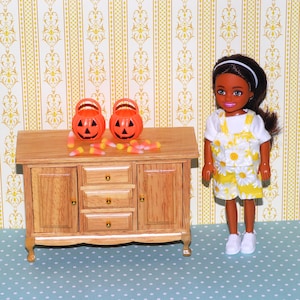 May include: A doll wearing a white shirt and yellow daisy print shorts and top. Two orange jack-o'-lantern buckets and candy corn sit on a wooden cabinet. The doll has dark hair and a white headband.
