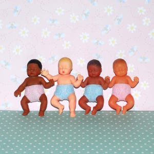 May include: Four small baby dolls with closed eyes and arms raised. The dolls have different skin tones and wear pastel blue and pink diapers. The background is a pink floral pattern.