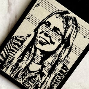 May include: A black rectangular tag with a scalloped top edge, featuring a black and white portrait of a smiling person. The portrait is overlaid on sheet music. A thin, gold-colored string is attached for hanging.