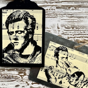 May include: Two black and white images of a man singing into a microphone. The images are printed on a tag with a string attached. The tag is on a white wooden surface. The text on the tag reads "mud, be my boy, his name was... shoes."