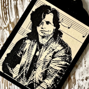 May include: Black and white image of a man with long hair wearing a jacket printed on a sheet of music. The image is printed on a black tag with a string attached.