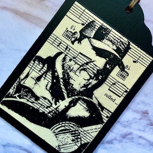 May include: Black and white image of a musician with a hat and beard singing into a microphone. The image is printed on a black tag with a white background. The tag has music notes and chords printed on it. The text "rolled" is visible on the tag.
