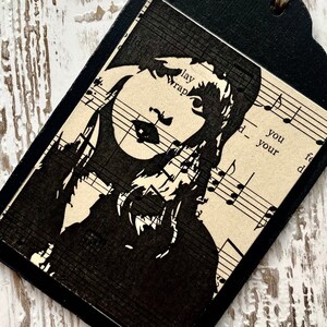 May include: Black and white silhouette of a woman with long hair on a vintage sheet music background. The music notes are visible in the background. The tag is black with a scalloped edge.