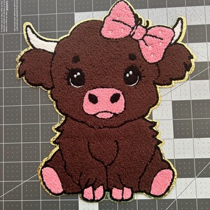 May include: A brown, textured patch in the shape of a Highland cow, featuring a pink bow, pink nose, and pink hooves. The patch has a gold border and large, expressive eyes. The cow is sitting down.