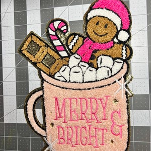 May include: A festive Christmas-themed patch featuring a gingerbread man in a pink hat and scarf, a candy cane, and a chocolate bar, all nestled in a pink mug filled with marshmallows. The mug reads "MERRY & BRIGHT" in pink lettering.