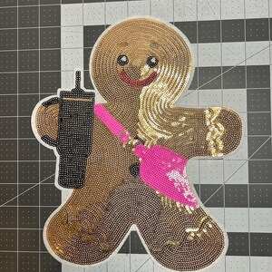 May include: A sequined gingerbread man patch with a black coffee cup and a pink broom. The gingerbread man is brown and gold, with a smiling face and black eyes. The patch is on a white background.