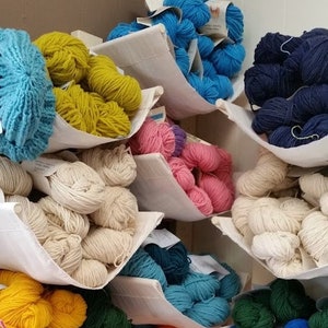 以下が含まれることがあります： 白い布製のバスケットに、さまざまな色の糸巻きが並べられています。糸巻きは色ごとに並べられており、青、黄、白、緑、オレンジ、赤などがあります。