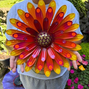 May include: A multi-layered metal flower wall art piece featuring elongated petals in a gradient of yellow, orange, and magenta. The center is a raised, dark circular dome decorated with a pattern of small, raised yellow dots.