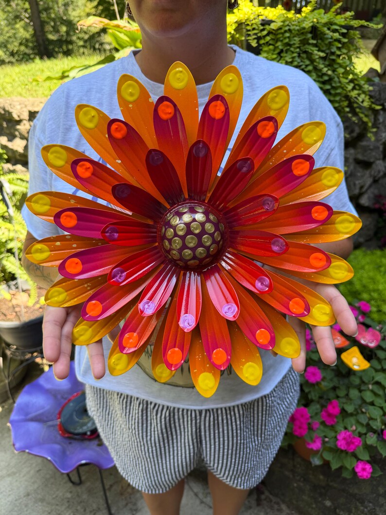 May include: A multi-layered metal flower wall art piece featuring elongated petals in a gradient of yellow, orange, and magenta. The center is a raised, dark circular dome decorated with a pattern of small, raised yellow dots.
