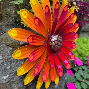 May include: A multi-layered metal flower wall art piece featuring elongated petals in vibrant shades of orange, red, and magenta. Each petal is accented with a raised yellow or pink dot. The center is a dark purple dome with gold studs.