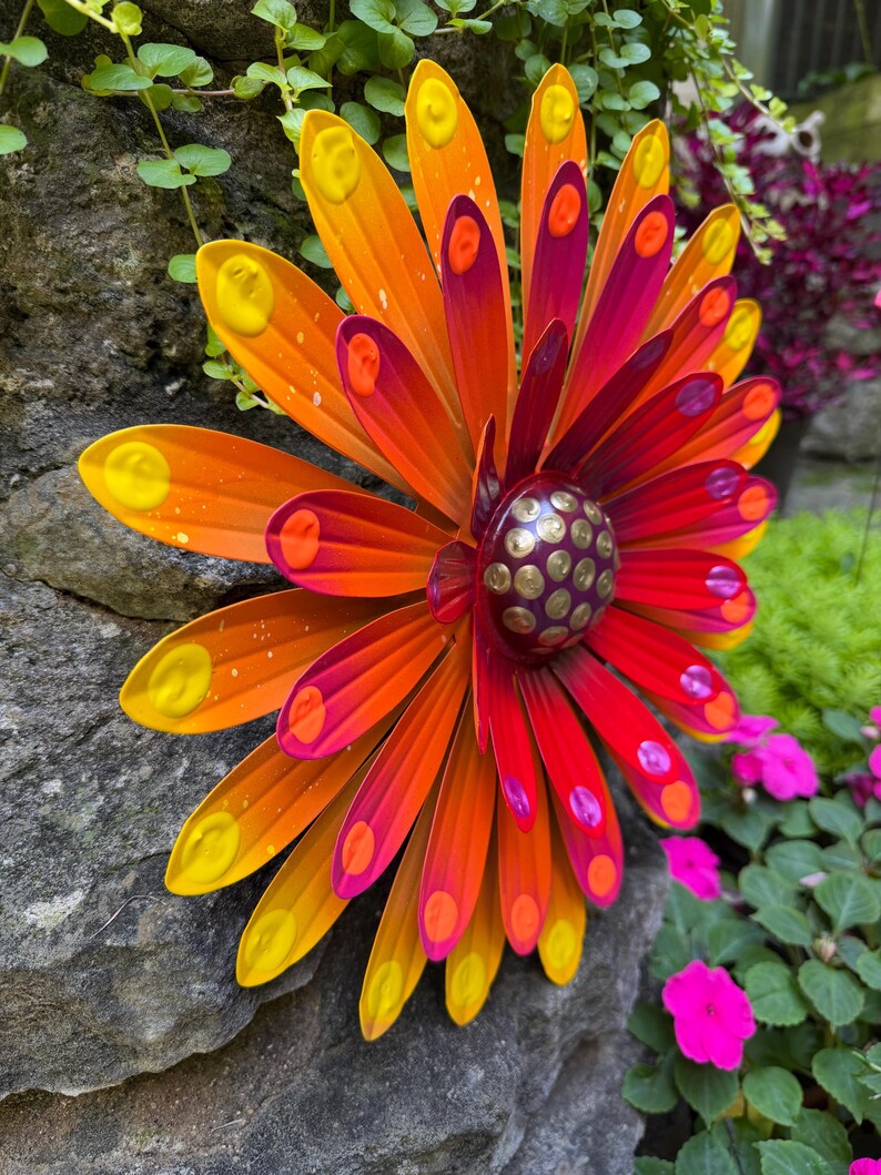 May include: A multi-layered metal flower wall art piece featuring elongated petals in vibrant shades of orange, red, and magenta. Each petal is accented with a raised yellow or pink dot. The center is a dark purple dome with gold studs.