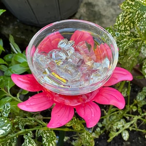 May include: A pink plastic flower with a clear glass bowl in the center filled with ice cubes. The flower is on a green metal stake and is surrounded by green plants.