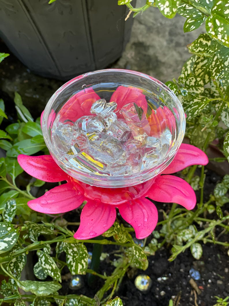May include: A pink plastic flower with a clear glass bowl in the center filled with ice cubes. The flower is on a green metal stake and is surrounded by green plants.