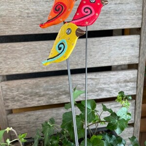 May include: Three colorful bird figurines with painted details on metal stems. The birds are yellow, orange, and red with black eyes and swirling patterns.