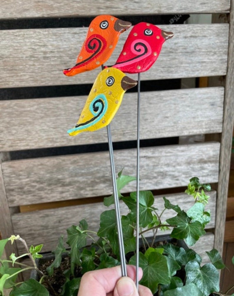 May include: Three colorful bird figurines with painted details on metal stems. The birds are yellow, orange, and red with black eyes and swirling patterns.