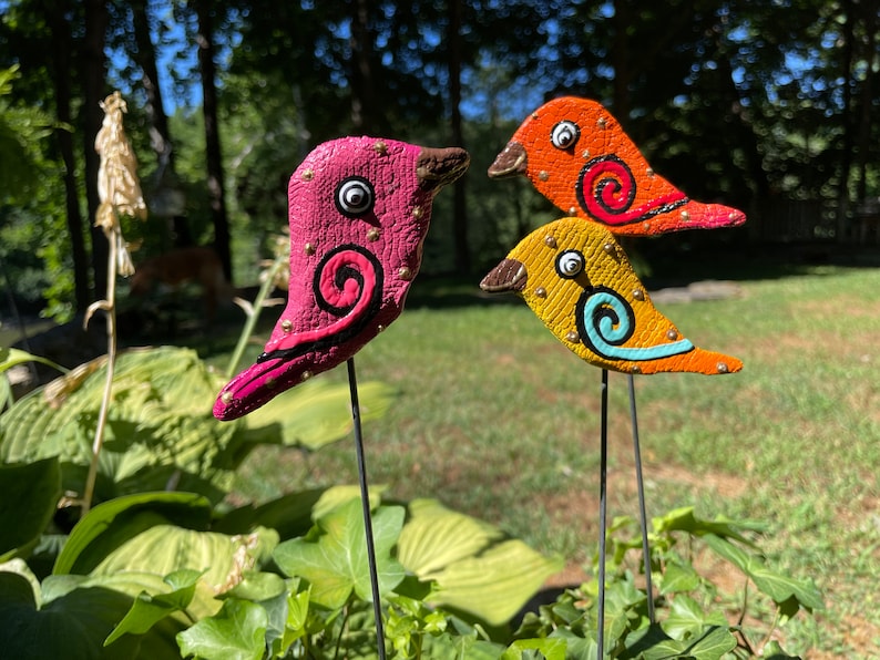 May include: Three colorful ceramic bird figurines with gold accents and painted details. The birds are on black metal stakes and are arranged in a row.