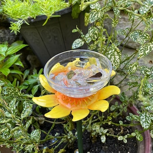 May include: A yellow and orange sunflower-shaped bird feeder with a clear glass bowl on top. The feeder is sitting in a pot of green plants.