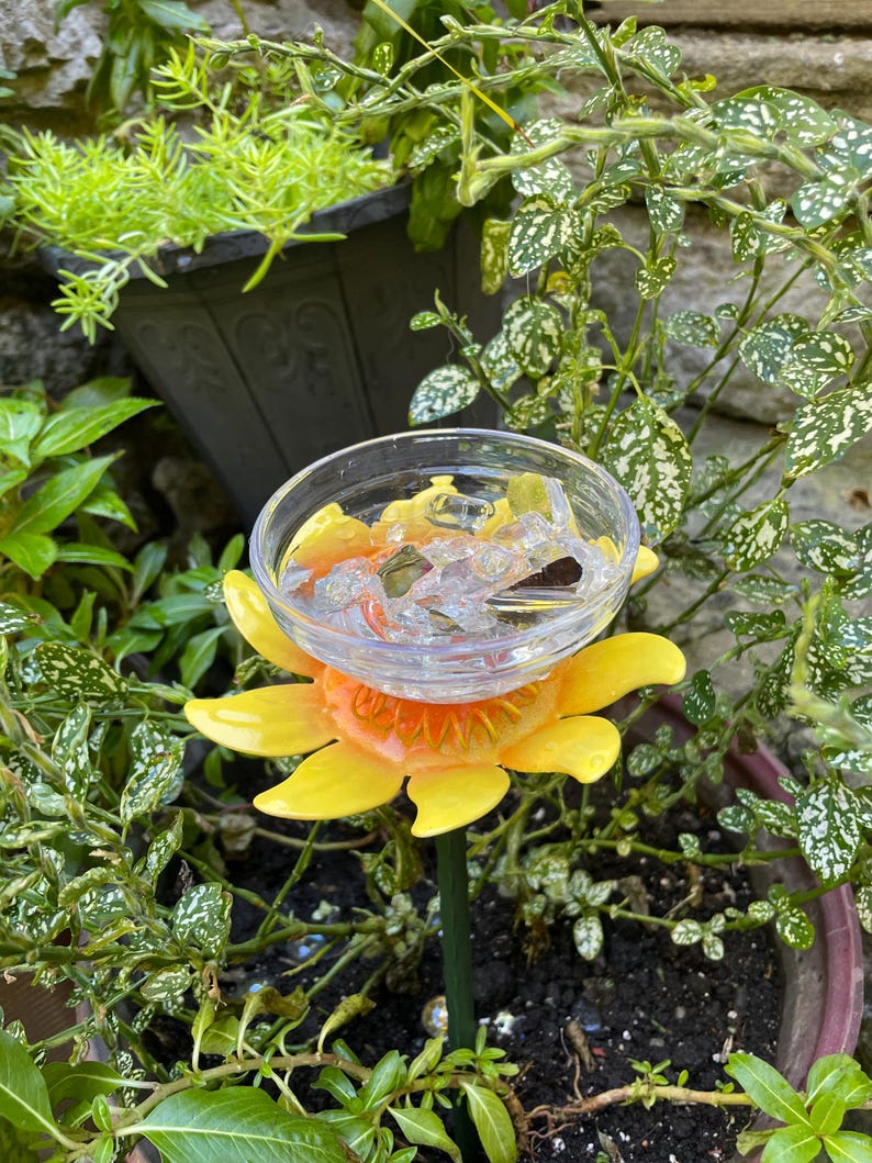 May include: A yellow and orange sunflower-shaped bird feeder with a clear glass bowl on top. The feeder is sitting in a pot of green plants.