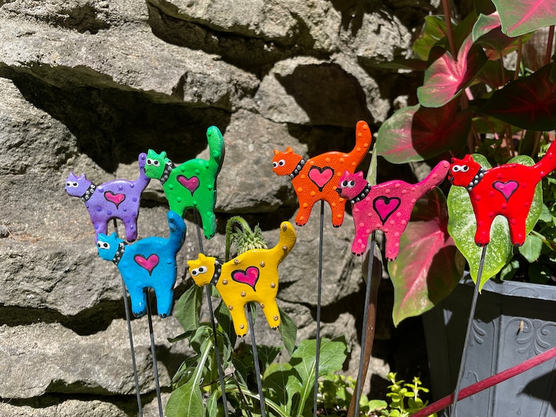 Puede incluir: Siete estacas de jard&iacute;n de gato de colores con corazones y lunares. Los gatos est&aacute;n hechos de arcilla pintada y est&aacute;n unidos a estacas de metal. Las estacas se insertan en el suelo.