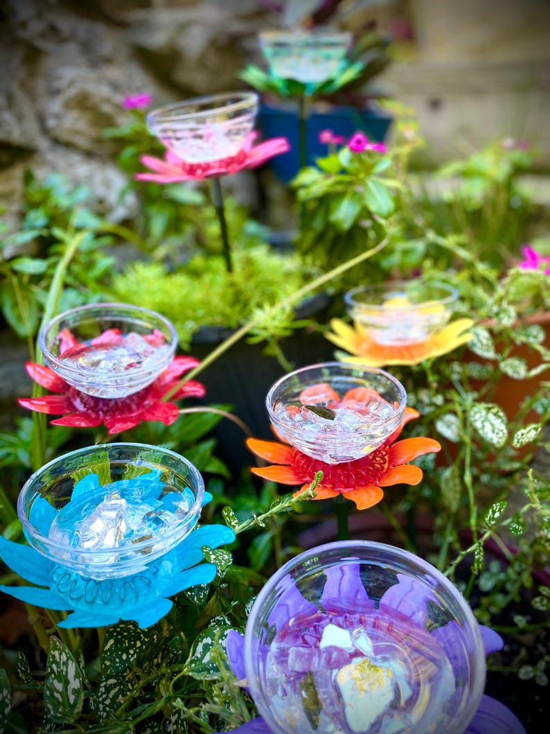 May include: Colorful plastic flowers with clear cups in the center, arranged in a garden setting. The flowers are in shades of pink, red, orange, blue, and purple.