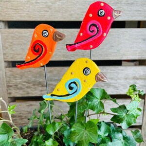May include: Three colorful ceramic bird figurines with painted details, each perched on a metal stake, are displayed in a potted plant. The birds are red, orange, and yellow with black and blue accents.